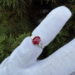 May include: A silver-toned ring featuring a central oval-cut red gemstone flanked by two smaller round-cut clear stones on each side. The ring is displayed on a white fabric-covered finger against a background of green pine needles.
