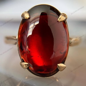 May include: A gold ring featuring a large, oval-shaped, deep red gemstone set in a four-prong mounting. The stone has a smooth, polished surface with visible internal reflections. The metal band is thin and polished, showing a warm gold tone.
