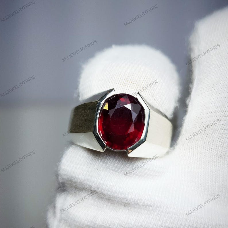 Pu&ograve; includere: Un anello d'argento con una grande pietra preziosa rosso scuro a taglio ovale. L'anello ha un design moderno e geometrico con una fascia larga e spalle angolari. La pietra preziosa &egrave; il punto focale, incastonata in un'incastonatura d'argento lucido.