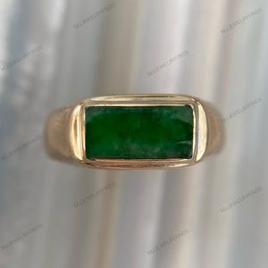 May include: A gold ring with a rectangular, emerald-green stone. The ring has a simple, classic design with a polished finish. The stone is set flush within the gold band, creating a sleek and modern look. The ring is a piece of jewelry.