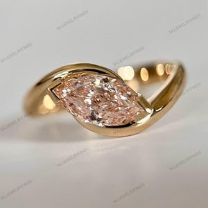 May include: A gold ring featuring a marquise-cut, light pink gemstone. The ring's band curves around the stone, creating a modern, elegant design. The ring is set against a soft, neutral background, highlighting the jewelry's details.