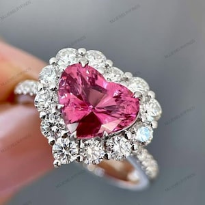 Puede incluir: Anillo con una piedra preciosa rosa en forma de corazón, rodeada por un halo de piedras preciosas más pequeñas y transparentes. La banda del anillo también está adornada con piedras preciosas transparentes, creando un efecto brillante. El anillo está engastado en metal plateado o blanco.