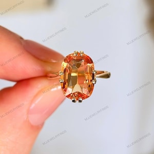 May include: A gold ring with a large, oval-cut, orange gemstone. The gemstone is the focal point, with a faceted surface that catches the light. The ring's band is a simple, thin gold band, and the gemstone is secured with decorative prongs.