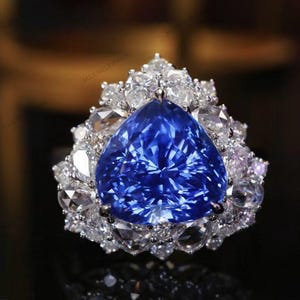 May include: A stunning ring featuring a large, triangular, deep blue gemstone as the centerpiece, surrounded by a halo of smaller, clear gemstones. The ring's design is intricate, with the gemstones set in a silver or white metal band.