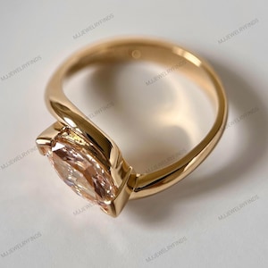 May include: A gold ring with a large, light peach-colored gemstone. The ring has a unique, modern design with a smooth, polished band that curves around the gemstone. The gemstone is set in a bezel setting.