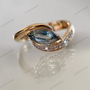 May include: A gold ring with a light blue, marquise-cut gemstone as the focal point. The band curves around the central stone, with small, clear gemstones set along one side. The ring is presented on a reflective surface.