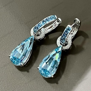 May include: A pair of elegant earrings featuring pear-shaped, light blue gemstones. The earrings have a silver-colored metal setting, with smaller blue stones and clear crystals accenting the hoops. The gemstones are faceted, catching the light.