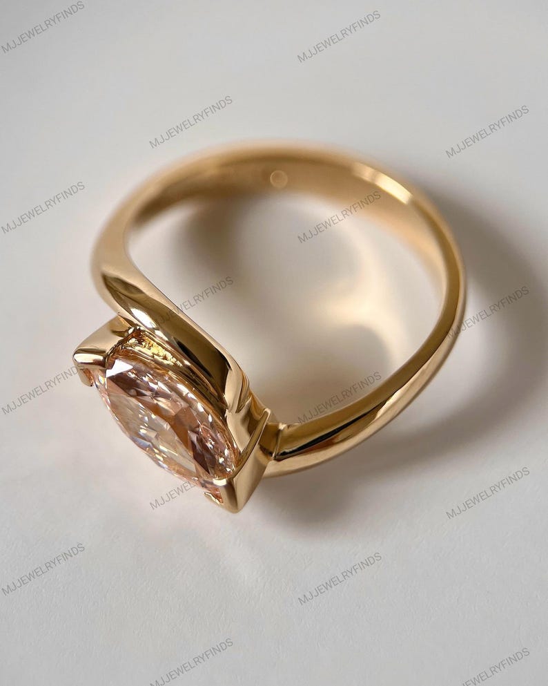 May include: A gold ring with a large, light peach-colored gemstone. The ring has a unique, modern design with a smooth, polished band that curves around the gemstone. The gemstone is set in a bezel setting.