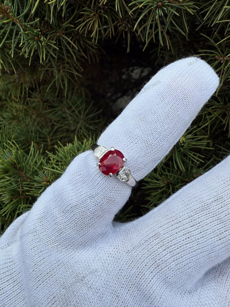 May include: A silver-toned ring featuring a central oval-cut red gemstone flanked by two smaller round-cut clear stones on each side. The ring is displayed on a white fabric-covered finger against a background of green pine needles.