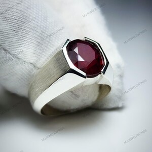 Pu&ograve; includere: Un anello d'argento con una grande pietra preziosa rosso scuro a taglio ovale. L'anello ha una fascia in metallo spazzolato e un'incastonatura in argento lucido. Un gioiello.