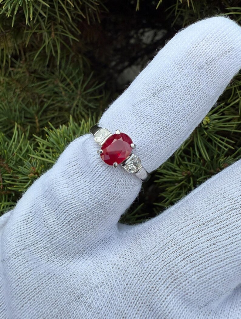 May include: A silver ring featuring a central oval-cut red ruby flanked by two smaller round-cut white diamonds on each side. The band is polished silver, showcasing a classic three-stone design with a vibrant gemstone centerpiece.