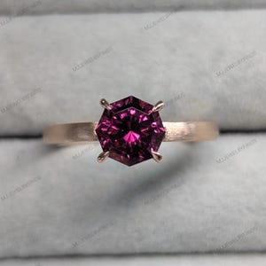 May include: A rose gold ring featuring a faceted, hexagonal, deep magenta gemstone. The ring has a simple band and four prongs securing the stone. The ring is set against a soft gray background.