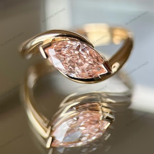 May include: A gold ring featuring a marquise-cut, light pink gemstone. The ring's band curves around the stone, creating a modern, elegant design. The ring is displayed on a reflective surface, enhancing its visual appeal.