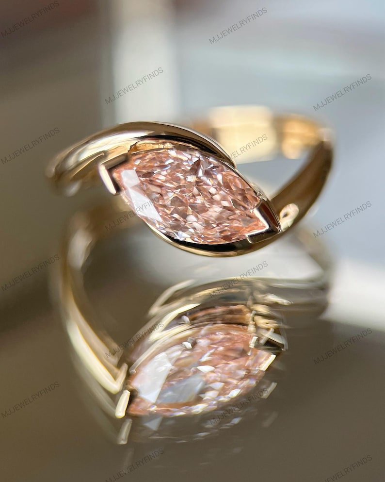 May include: A gold ring featuring a marquise-cut, light pink gemstone. The ring's band curves around the stone, creating a modern, elegant design. The ring is displayed on a reflective surface, enhancing its visual appeal.