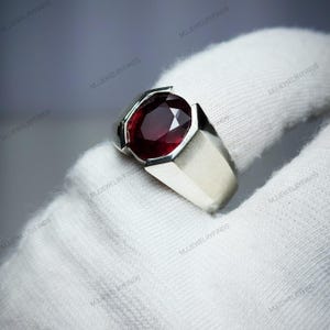 Pu&ograve; includere: Un anello d'argento con una grande pietra preziosa rosso scuro a taglio ovale. L'anello ha un design semplice e moderno con una fascia rettangolare e un'incastonatura a castone. La pietra preziosa &egrave; il punto focale, catturando la luce e mostrando il suo colore intenso.
