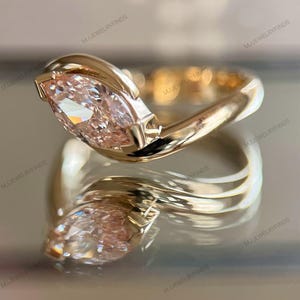 May include: A gold ring with a marquise-cut, light pink gemstone. The ring's band curves around the stone, creating a modern, elegant design. The ring is displayed on a reflective surface, showcasing its intricate details. The text "MJJEWELRYFINDS" is visible.
