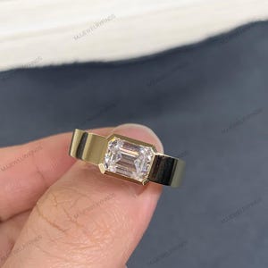 May include: A gold ring with a rectangular, clear gemstone. The ring has a minimalist design with a wide band. The gemstone is set in a bezel setting. The ring's design is modern and elegant.