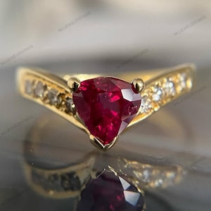 May include: A gold ring featuring a central trillion-cut deep red gemstone held by three prongs. The band is V-shaped and accented with small, clear, round-cut stones set along both sides of the shank, leading up to the central red stone.