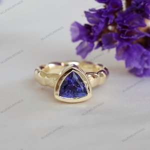 May include: A gold ring with a triangular, deep blue gemstone. The ring has a textured band and is set against a soft, white background with purple flowers in the background. The ring is a piece of jewelry.