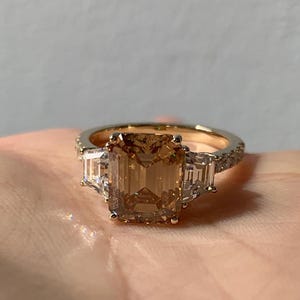 Puede incluir: Un anillo de oro con una gran gema rectangular de color marrón claro como pieza central. Dos gemas claras rectangulares más pequeñas flanquean la piedra principal. La banda del anillo también está adornada con gemas claras más pequeñas, creando un efecto brillante.