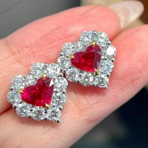May include: A pair of heart-shaped earrings. Each earring has a vibrant red gemstone centre, surrounded by a halo of sparkling white gemstones. The earrings have a gold-coloured setting. The image is a close-up of the earrings.