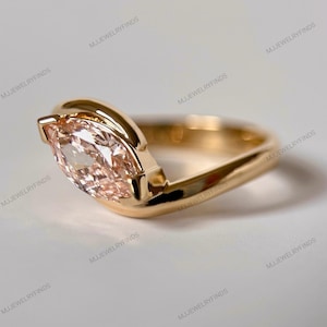 May include: A gold ring featuring a marquise-cut, peach-colored gemstone. The ring's band has a unique, curved design that wraps around the stone. The ring is set against a white background, highlighting its elegant simplicity. The ring is a piece of jewelry.