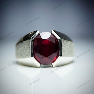May include: A silver ring featuring a large, oval-cut, deep red gemstone. The ring has a brushed metal finish on the sides and a polished top. The gemstone is the focal point, reflecting light.