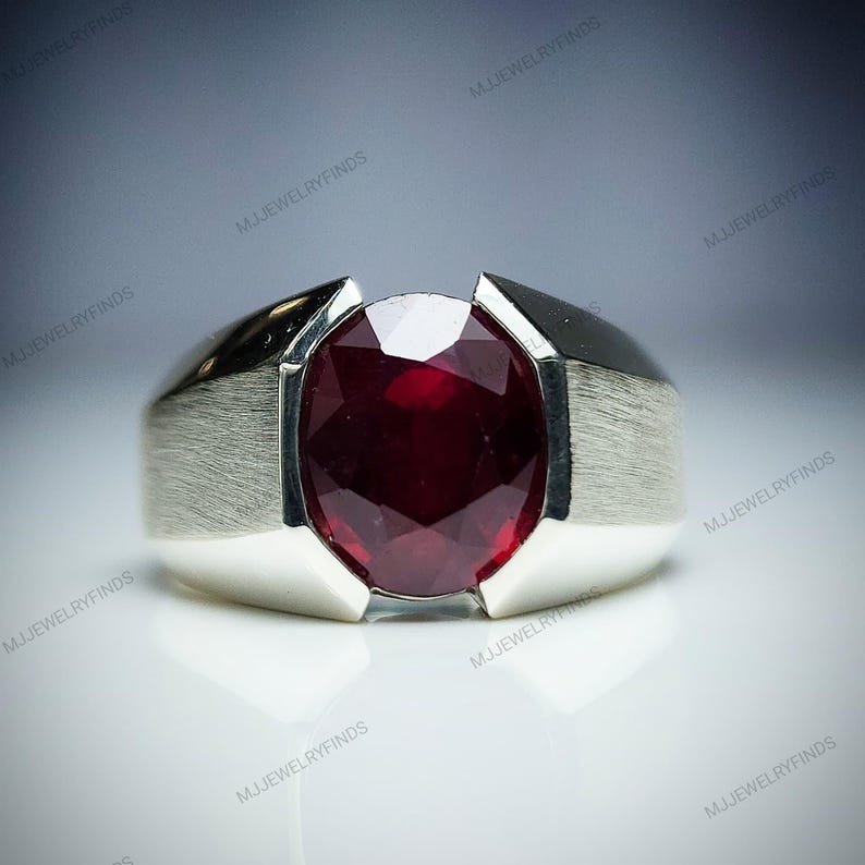 Pu&ograve; includere: Un anello d'argento con una grande pietra preziosa rosso scuro a taglio ovale. L'anello ha una finitura in metallo spazzolato sui lati e una parte superiore lucida. La pietra preziosa &egrave; il punto focale, che riflette la luce.