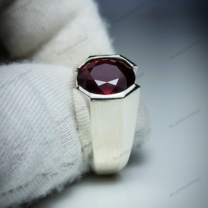 Pu&ograve; includere: Un anello d'argento con una grande pietra preziosa rosso scuro a taglio ovale. La pietra preziosa &egrave; incastonata in una lunetta e l'anello ha una fascia geometrica rettangolare. L'anello &egrave; mostrato su sfondo bianco.
