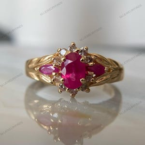 May include: A gold ring featuring a central oval-cut red ruby surrounded by a halo of small clear stones. Two marquise-cut red rubies are set on the sides of the central stone. The gold band has a textured, leaf-like pattern along the shank.
