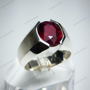 Pu&ograve; includere: Un anello d'argento con una grande pietra preziosa rosso scuro a taglio ovale. L'anello ha una finitura in metallo spazzolato sulla fascia e un'incastonatura angolare moderna che mette in risalto la brillantezza della gemma. L'anello &egrave; esposto su un supporto trasparente.