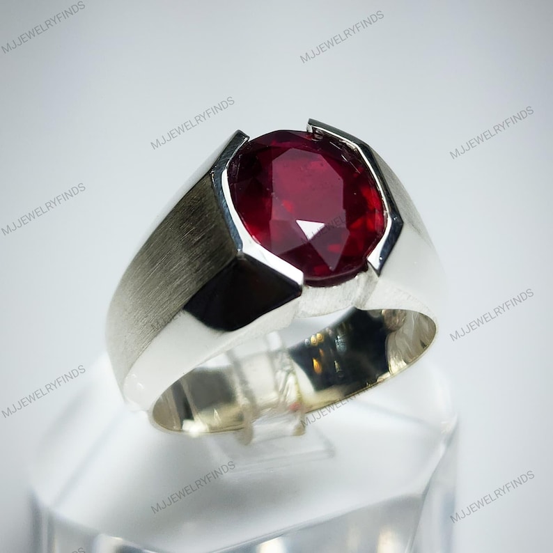 Pu&ograve; includere: Un anello d'argento con una grande pietra preziosa rosso scuro a taglio ovale. L'anello ha una finitura in metallo spazzolato sulla fascia e un'incastonatura angolare moderna che mette in risalto la brillantezza della gemma. L'anello &egrave; esposto su un supporto trasparente.