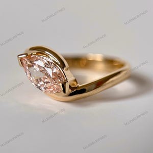 May include: A gold ring featuring a marquise-cut, peach-colored gemstone. The ring's band has a unique, curved design that cradles the gemstone. The ring is set against a neutral background, highlighting its elegant simplicity. The ring is a piece of jewelry.