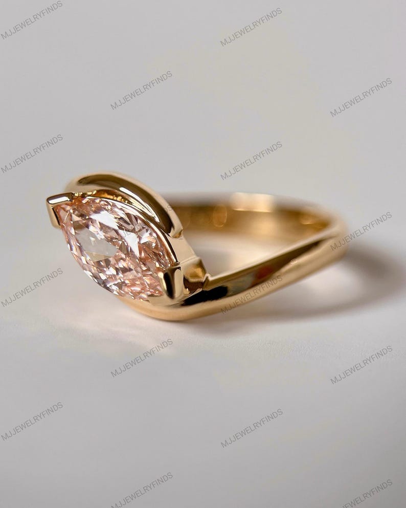May include: A gold ring featuring a marquise-cut, peach-colored gemstone. The ring's band has a unique, curved design that cradles the gemstone. The ring is set against a neutral background, highlighting its elegant simplicity. The ring is a piece of jewelry.