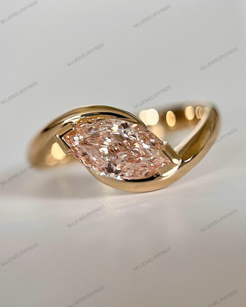 May include: A gold ring featuring a marquise-cut, light pink gemstone. The ring's band curves around the stone, creating a modern, elegant design. The ring is set against a soft, neutral background, highlighting the jewelry's details.