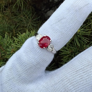 May include: A silver ring featuring a central oval-cut red ruby flanked by two smaller round-cut white diamonds on each side. The band is polished silver, showcasing a classic three-stone design with a vibrant gemstone centerpiece.