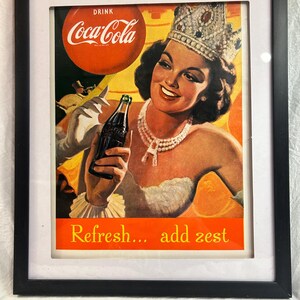 May include: Framed vintage Coca-Cola advertisement featuring a woman wearing a crown and holding a bottle of Coke. The ad has a red and orange color scheme with the text "Drink Coca-Cola" and "Refresh... add zest". The frame is black.