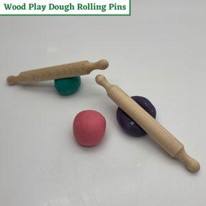 May include: Two wooden rolling pins for play dough, one resting on a green ball of dough and the other on a purple ball of dough. There is also a pink ball of dough.