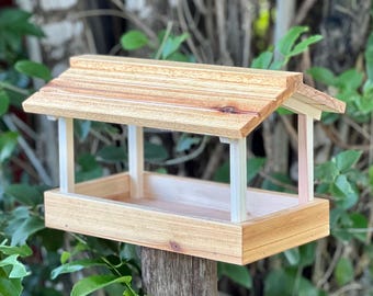 Puede incluir: Un comedero para pájaros de madera con una base rectangular, cuatro postes de soporte verticales blancos y un techo inclinado y sobresaliente hecho de tablones de madera de color marrón claro natural con vetas y nudos visibles.