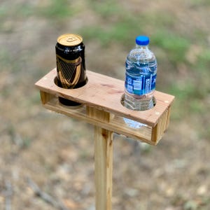 May include: A wooden drink holder with two circular cutouts, one holding a Guinness can and the other a water bottle. The holder is mounted on a wooden post, perfect for outdoor use. The Guinness can is black with gold lettering.