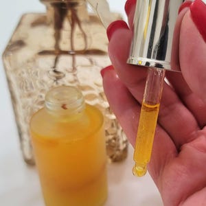 May include: A clear glass bottle filled with a yellow liquid, with a dropper held above it. The dropper is silver and glass, with a drop of the liquid suspended from the tip. A second bottle is in the background, along with a decorative vase.