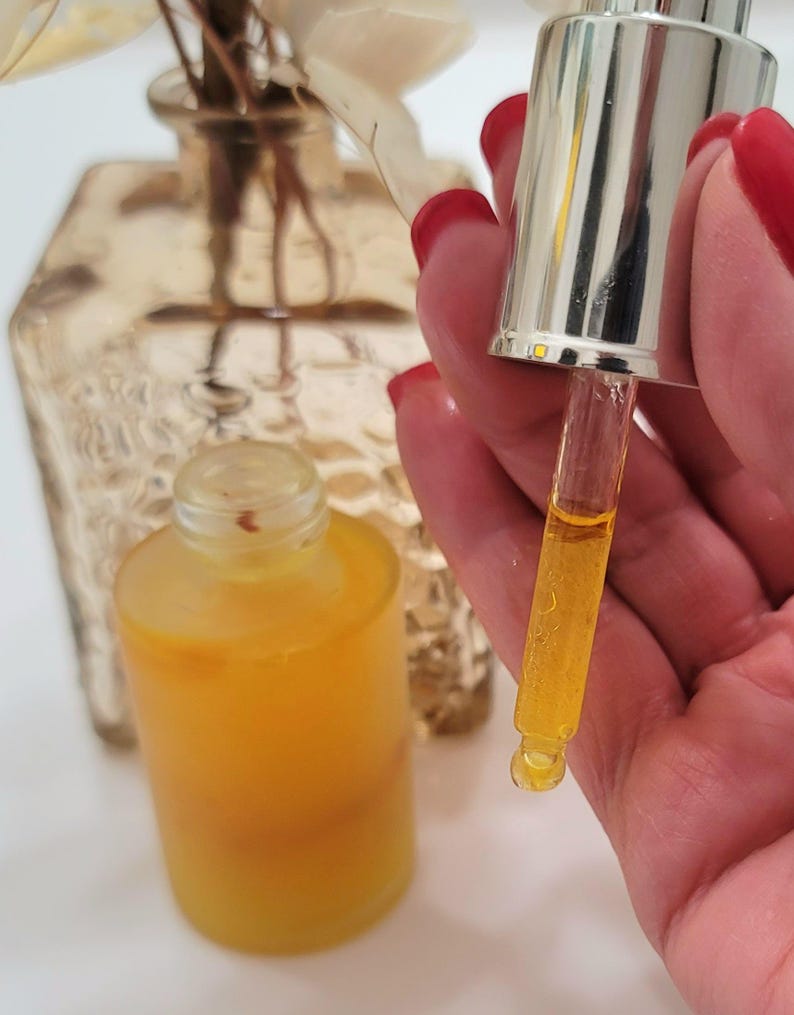 May include: A clear glass bottle filled with a yellow liquid, with a dropper held above it. The dropper is silver and glass, with a drop of the liquid suspended from the tip. A second bottle is in the background, along with a decorative vase.