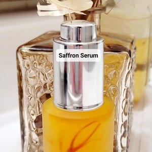 May include: A clear glass bottle filled with a yellow liquid, labeled "Saffron Serum." The bottle has a silver cap and is set against a blurred background of decorative glass and dried flowers. The serum appears to have saffron strands.