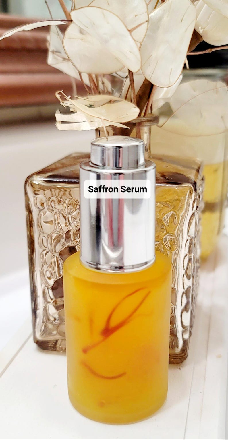 May include: A clear glass bottle filled with a yellow liquid, labeled "Saffron Serum." The bottle has a silver cap and is set against a blurred background of decorative glass and dried flowers. The serum appears to have saffron strands.