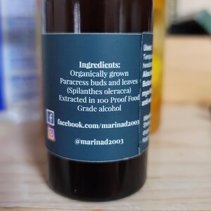 Może przedstawiać: Ciemnobrązowa szklana butelka z czarną zakrętką i niebieską etykietą. Na etykiecie wymieniono składniki: organicznie uprawiane pąki i liście Paracress, ekstrahowane w 100-procentowym alkoholu spożywczym. Wymieniono r&oacute;wnież uchwyty w mediach społecznościowych.