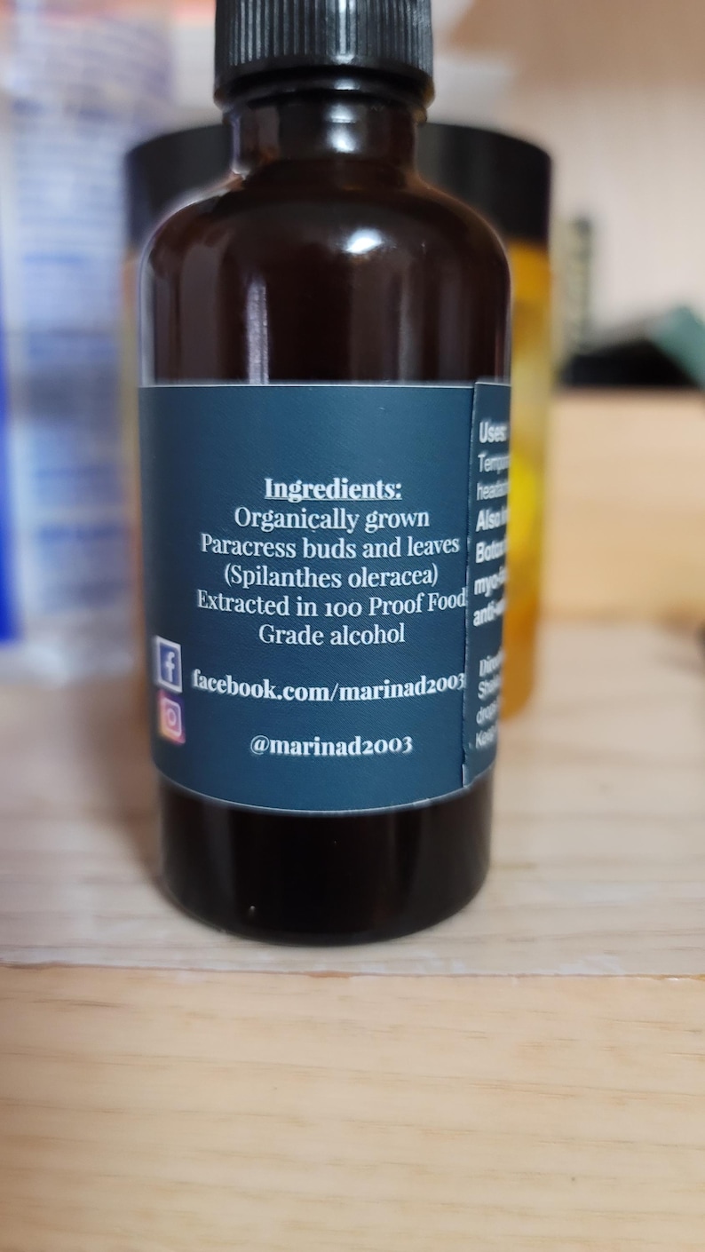 Może przedstawiać: Ciemnobrązowa szklana butelka z czarną zakrętką i niebieską etykietą. Na etykiecie wymieniono składniki: organicznie uprawiane pąki i liście Paracress, ekstrahowane w 100-procentowym alkoholu spożywczym. Wymieniono r&oacute;wnież uchwyty w mediach społecznościowych.
