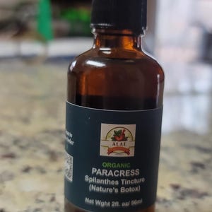 Może przedstawiać: Brązowa szklana butelka z czarną zakrętką, z etykietą "ORGANIC PARACRESS Spilanthes Tincture (Nature's Botox)". Etykieta zawiera nazwę marki "ALAE" i wagę netto 56 ml.
