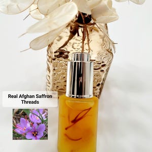 May include: A small, clear glass bottle with a silver cap, filled with a yellow liquid and saffron threads. The text "Real Afghan Saffron Threads" is displayed next to a photo of purple saffron flowers. A decorative vase with dried flowers is in the background.