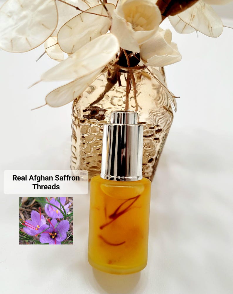 May include: A small, clear glass bottle with a silver cap, filled with a yellow liquid and saffron threads. The text "Real Afghan Saffron Threads" is displayed next to a photo of purple saffron flowers. A decorative vase with dried flowers is in the background.