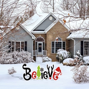 May include: A snowy winter scene with a house in the background. In the foreground, a large, colorful "Believe" sign is displayed on the lawn. The sign features a green Grinch character, a red Santa hat, and a snowflake.